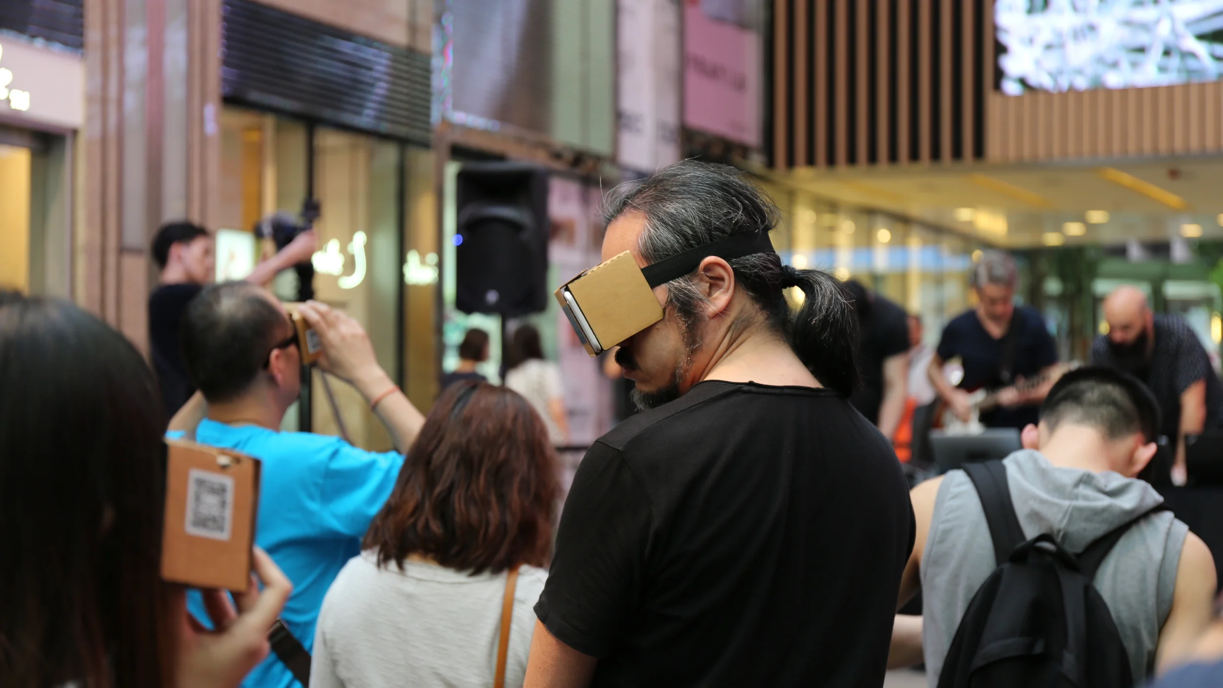 Audience members watch through cardboard VR viewers in a covered public space.