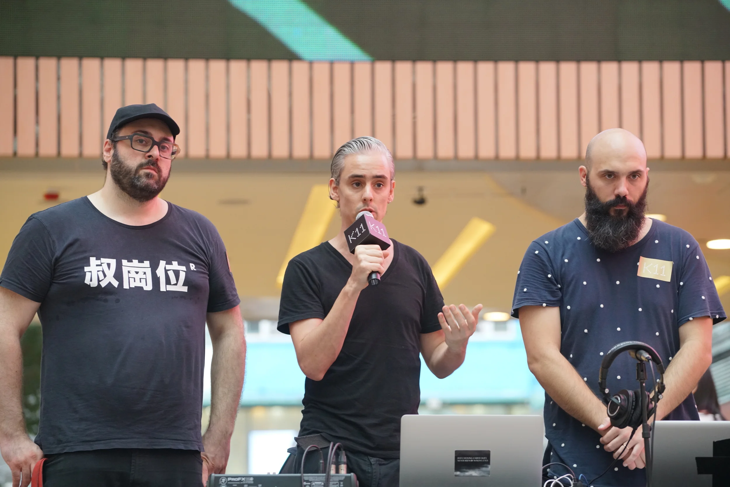 Three performers stand at a laptop while one speaks into a microphone.