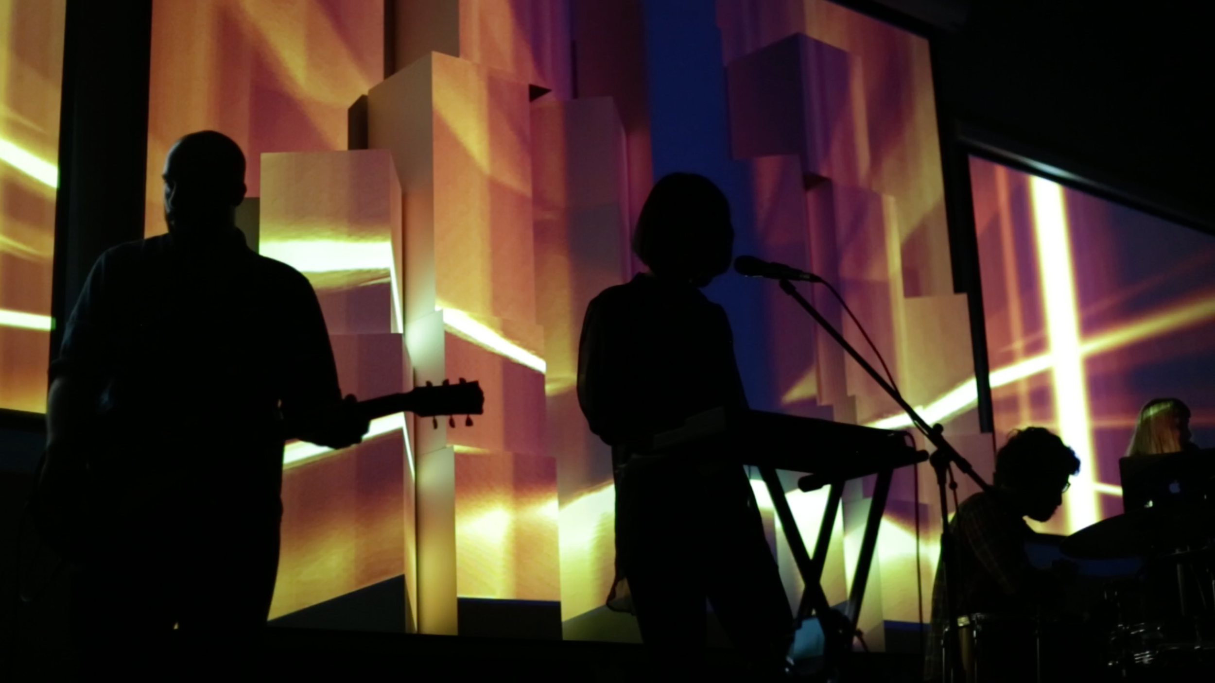 Musicians silhouetted with guitar and keyboard against a multicoloured geometric projection.