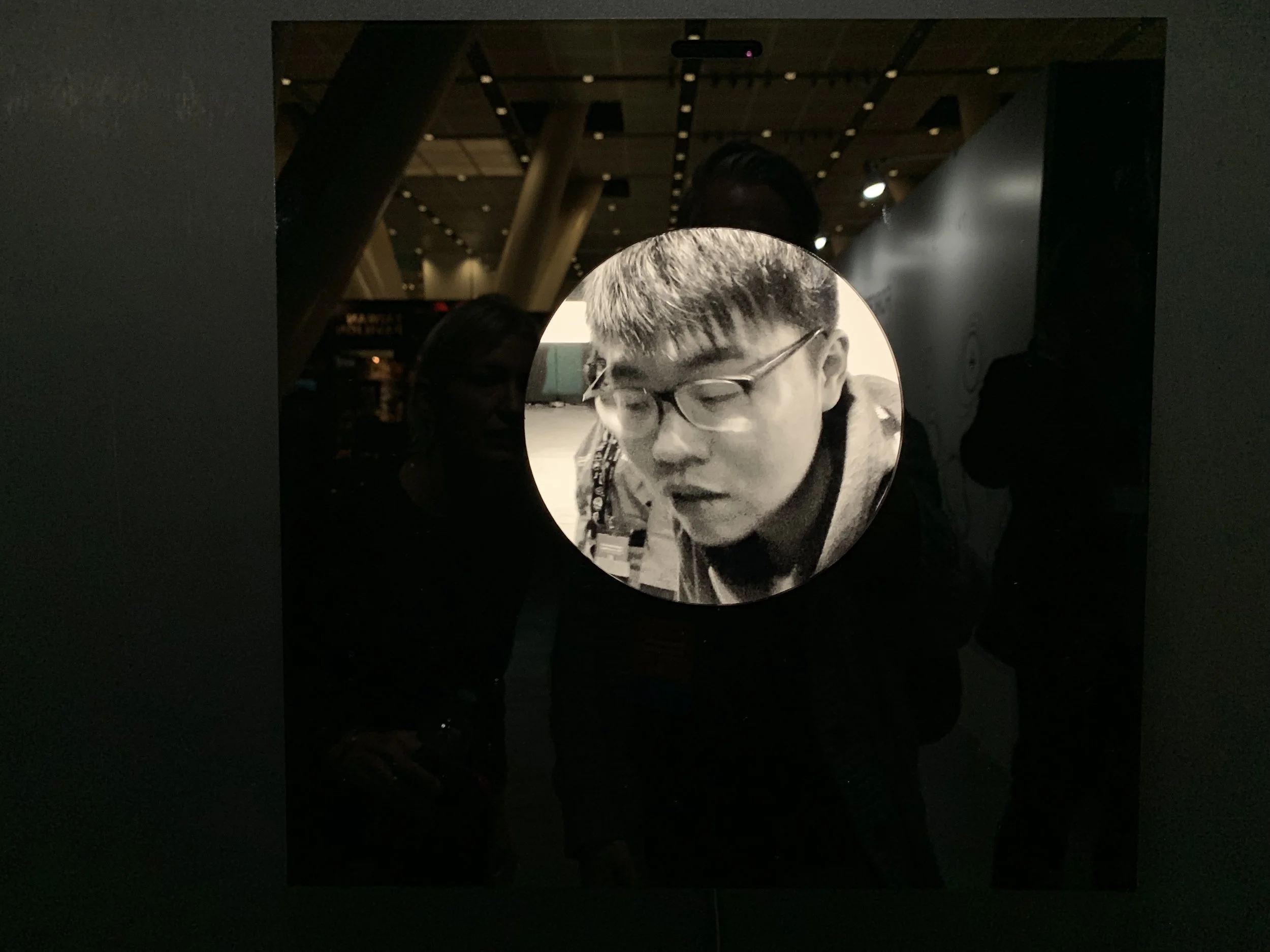A closed-eye portrait wearing glasses, surrounded by the reflection of the exhibition hall.