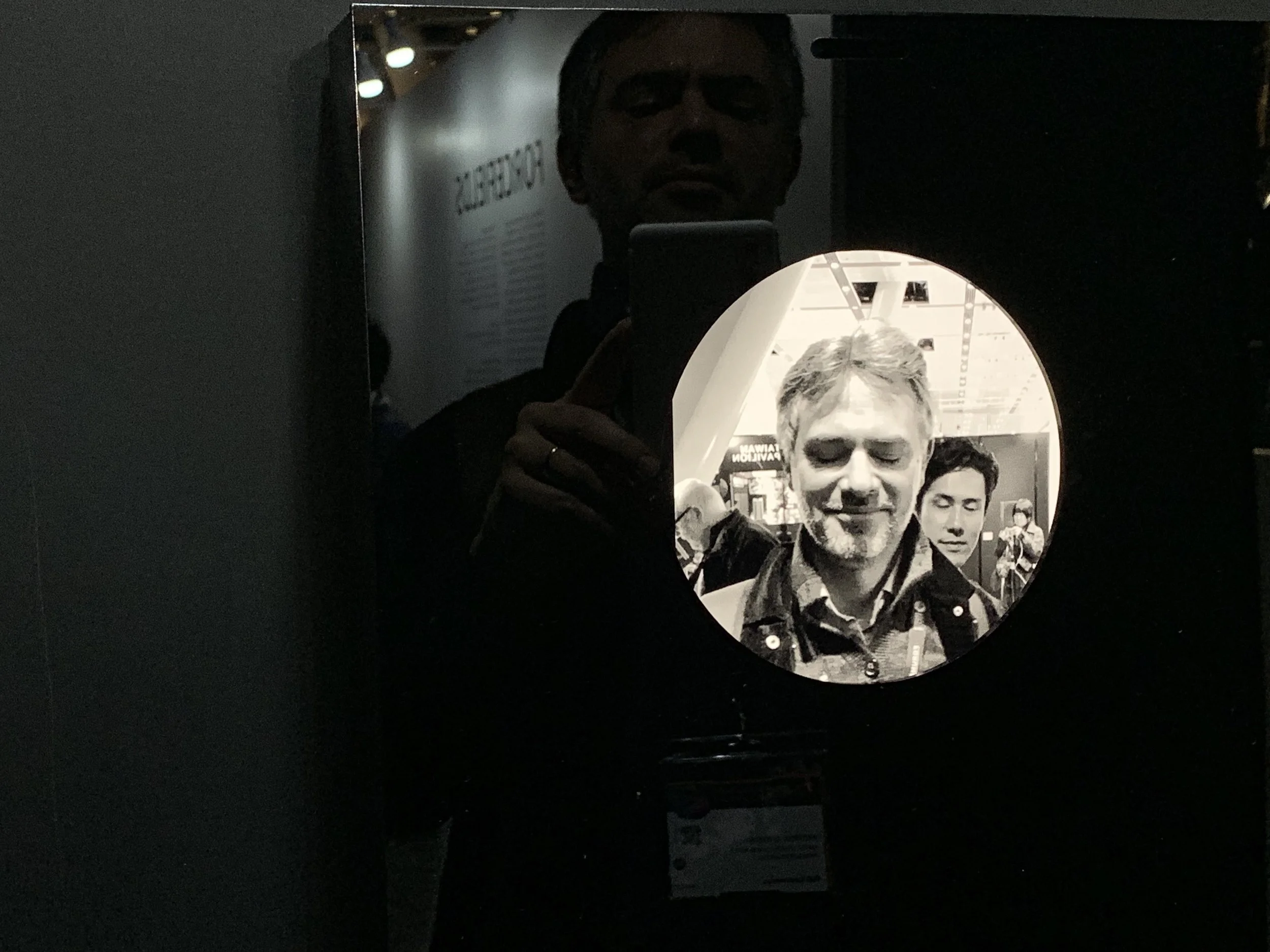 A smiling portrait in the circular display, with a reflected visitor holding a camera.